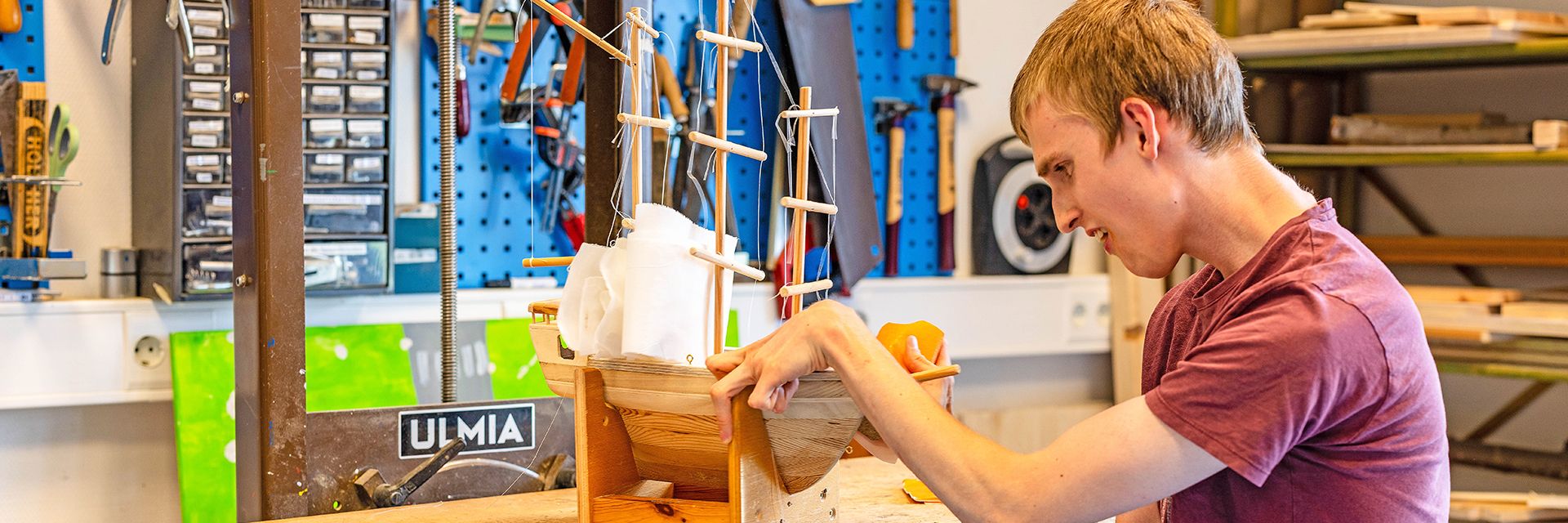 Ein junger Mann mit Behinderung sitzt in einer Werkstatt und arbeitet mit Schleifpapier an einem Modellschiff auf Holz.
