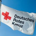 Im Wind weht eine weiße Flagge. Sie zeigt ein rotes Kreuz. Daneben steht in schwarzen Buchstaben: Deutsches Rotes Kreuz.