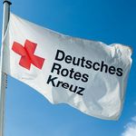 Im Wind weht eine weiße Flagge. Sie zeigt ein rotes Kreuz. Daneben steht in schwarzen Buchstaben: Deutsches Rotes Kreuz.